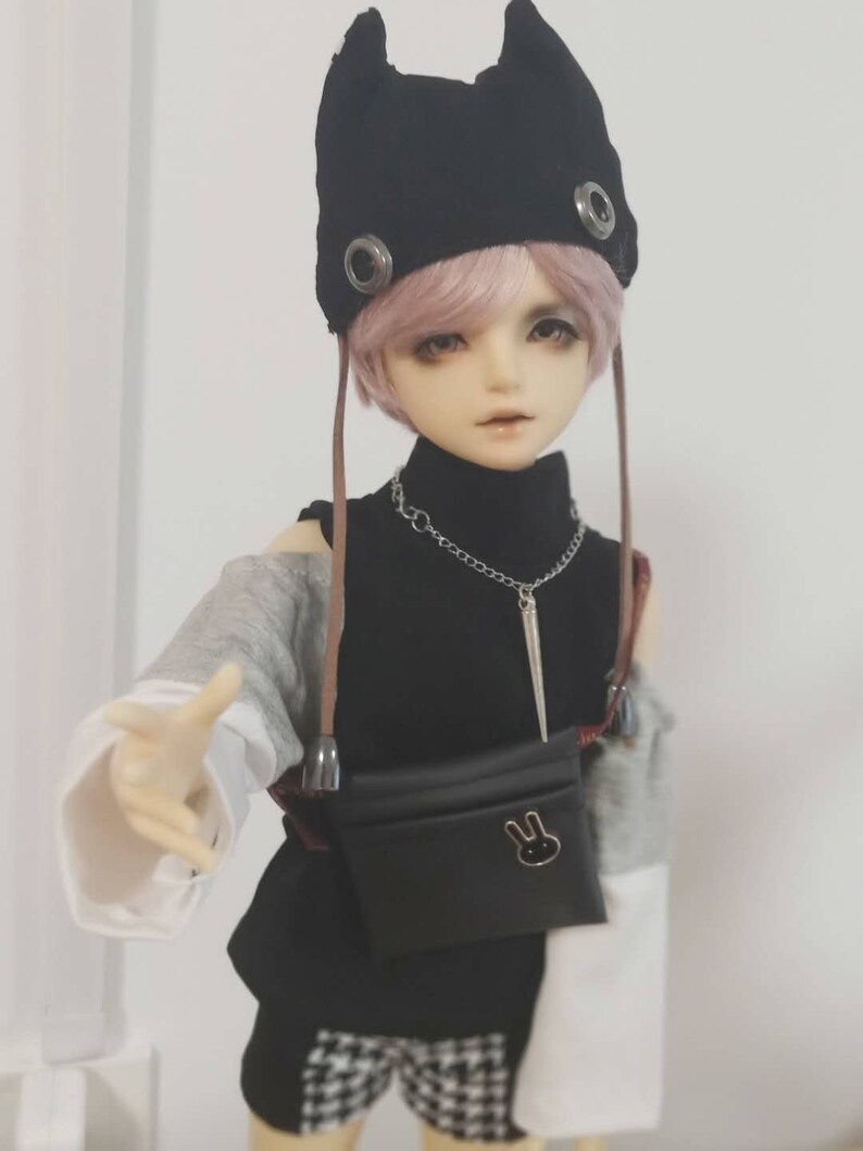 May include: A doll with pink hair wearing a black hat with ear flaps, a black turtleneck, a gray and white long-sleeved shirt, black shorts with a white and black checkered pattern, and a black fanny pack with a bunny design.