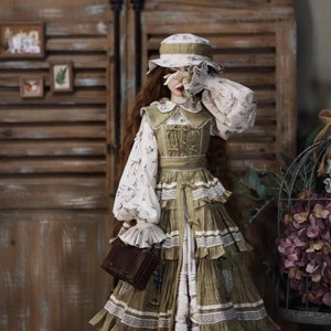 May include: A doll wearing a green and white dress with a floral print and a brown hat. The doll is holding a brown purse.