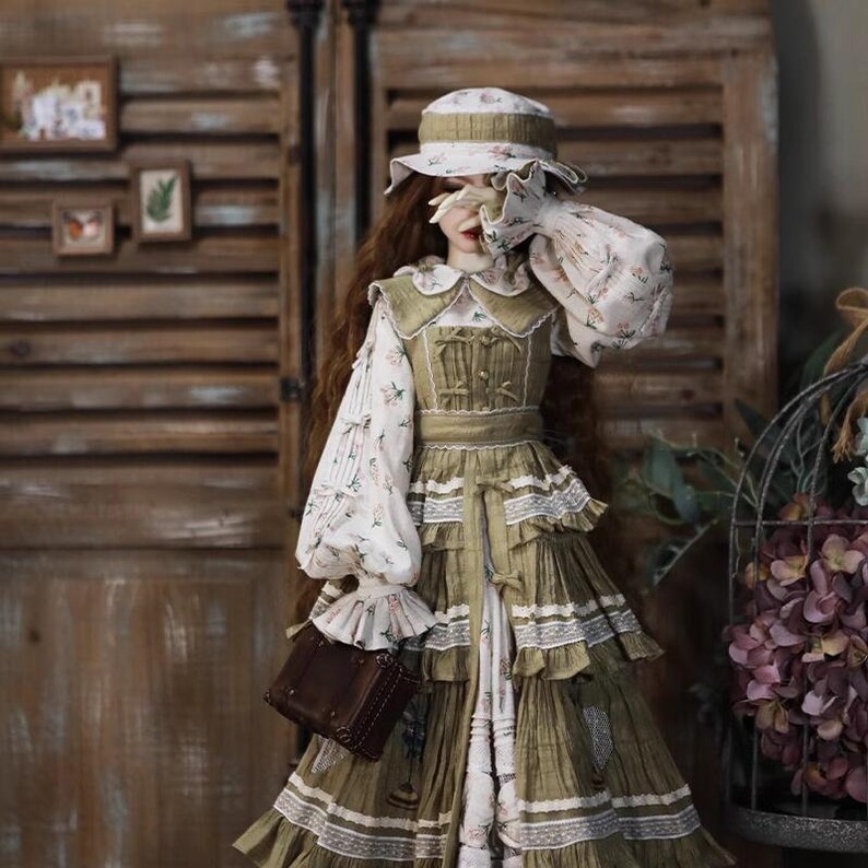 May include: A doll wearing a green and white dress with a floral print and a brown hat. The doll is holding a brown purse.