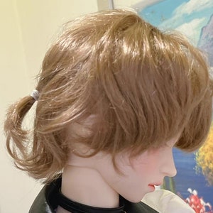 May include: A doll with a light brown wig styled in a short, layered cut with a ponytail secured with a white elastic band. The wig has a natural, slightly wavy texture.