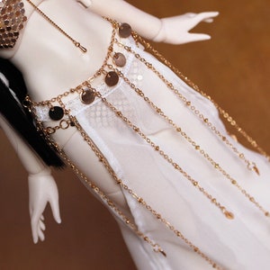 May include: A white sheer skirt with a gold chain belt featuring round charms and small bells. The skirt is made of a delicate fabric and has a flowing silhouette.