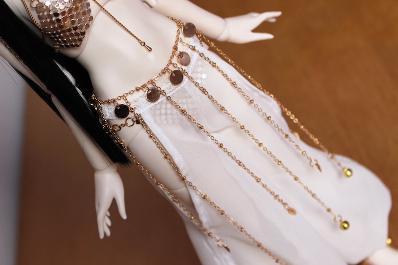 May include: A white sheer skirt with a gold chain belt featuring round charms and small bells. The skirt is made of a delicate fabric and has a flowing silhouette.