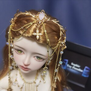 May include: A gold-toned headpiece featuring delicate metal leaf accents, multiple dangling chains, and a central pink teardrop-shaped stone. The accessory is adorned with small clear crystals and a cross-shaped charm, resting on a doll with light brown hair.