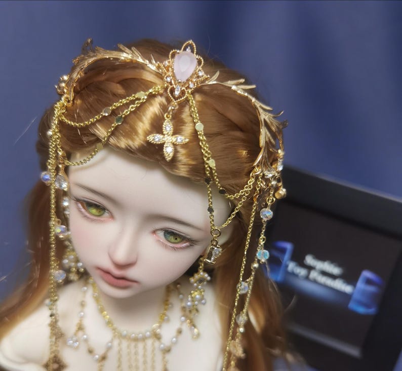 May include: A gold-toned headpiece featuring delicate metal leaf accents, multiple dangling chains, and a central pink teardrop-shaped stone. The accessory is adorned with small clear crystals and a cross-shaped charm, resting on a doll with light brown hair.