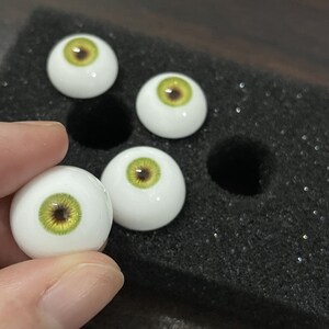 May include: Four white doll eyes with green and brown irises. The eyes are displayed on a black foam surface.