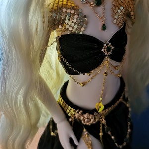 May include: A doll wearing a black and gold costume with green gem accents. The costume includes a chainmail top, a black bra with gold chains, a gold belt, and a black skirt. The doll has long blonde hair.
