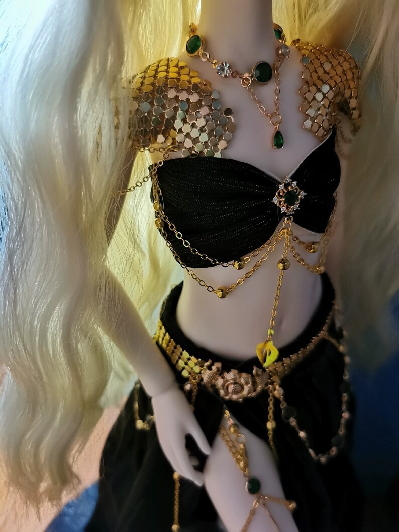 May include: A doll wearing a black and gold costume with green gem accents. The costume includes a chainmail top, a black bra with gold chains, a gold belt, and a black skirt. The doll has long blonde hair.