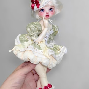 May include: A detailed doll with a white wig and blue eyes, wearing a green and cream dress with a matching hat. The doll's hat is adorned with a red bow and a decorative red fruit. The doll is wearing white shoes with a small heel.