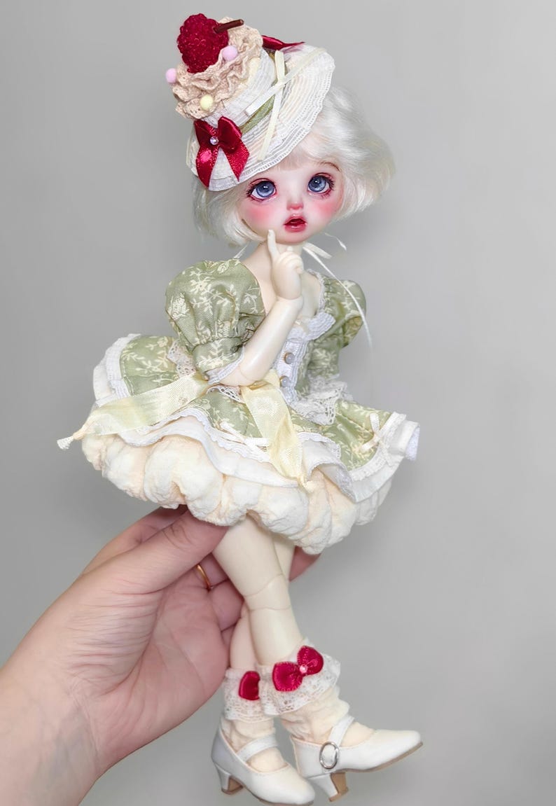 May include: A detailed doll with a white wig and blue eyes, wearing a green and cream dress with a matching hat. The doll's hat is adorned with a red bow and a decorative red fruit. The doll is wearing white shoes with a small heel.