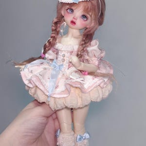 May include: A doll with pink hair in braids, wearing a pink dress with a floral pattern, a cupcake-shaped hat, and white shoes. The dress has a ruffled skirt and blue ribbon accents. The doll is held by a hand.