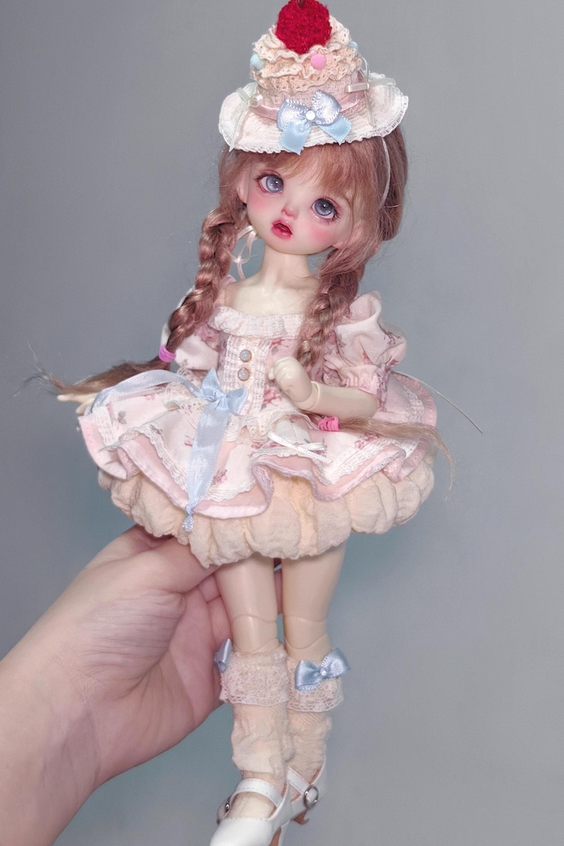 May include: A doll with pink hair in braids, wearing a pink dress with a floral pattern, a cupcake-shaped hat, and white shoes. The dress has a ruffled skirt and blue ribbon accents. The doll is held by a hand.