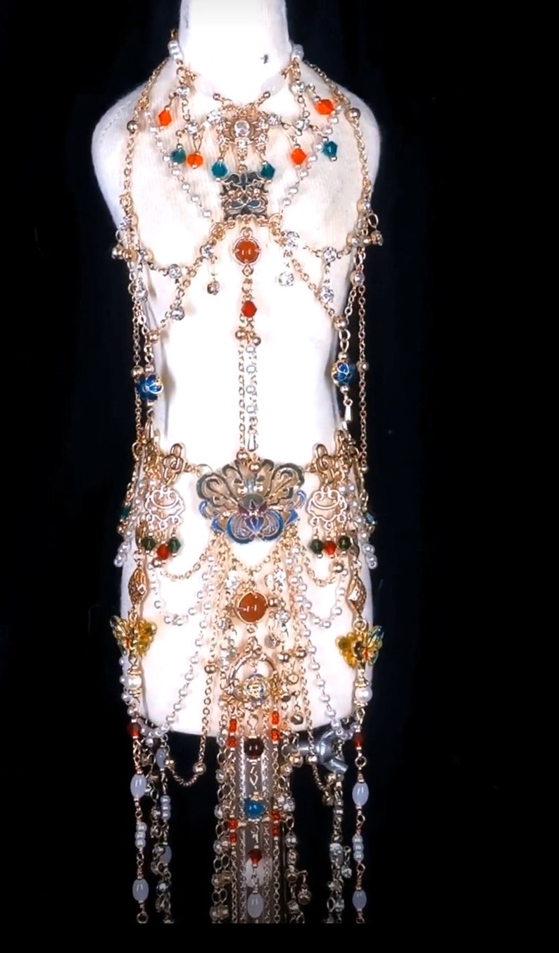 May include: A gold and silver body chain with multiple layers of beads and charms. The chain features a large, ornate pendant with blue and gold enamel. The beads are red, orange, green, and white.