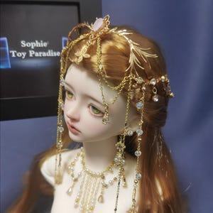 May include: A doll with long reddish-brown hair wearing a gold-toned headpiece featuring delicate metal branches, hanging chains, and clear crystals. A matching gold-toned necklace with white pearls and dangling crystal accents adorns the neck.