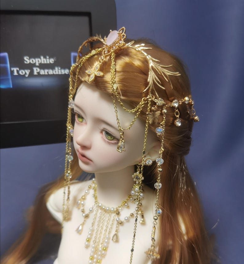May include: A doll with long reddish-brown hair wearing a gold-toned headpiece featuring delicate metal branches, hanging chains, and clear crystals. A matching gold-toned necklace with white pearls and dangling crystal accents adorns the neck.