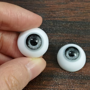 May include: Two white doll eyes with black pupils and silver glitter accents. The eyes are on a wooden surface.