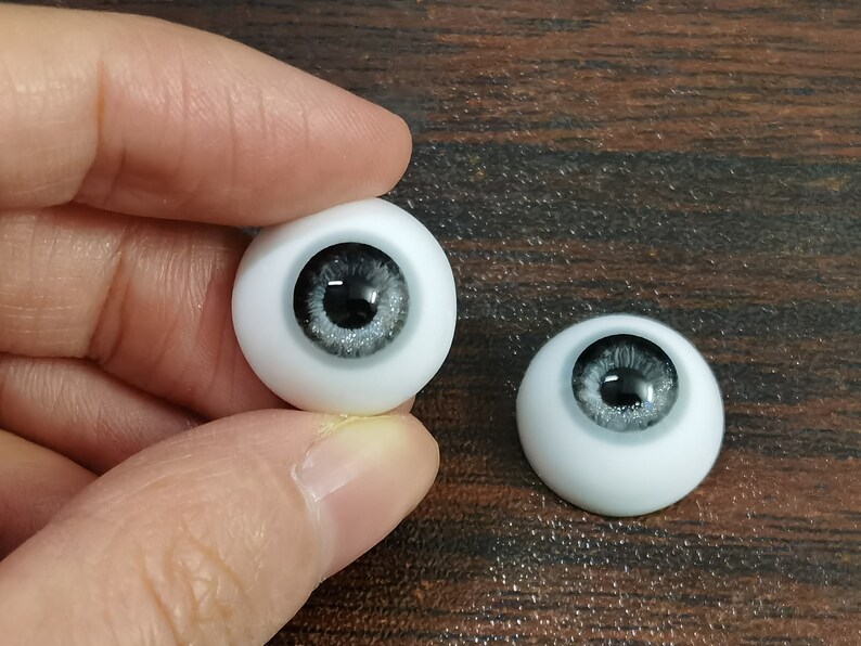 May include: Two white doll eyes with black pupils and silver glitter accents. The eyes are on a wooden surface.