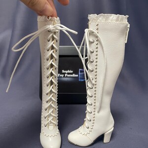 Pode incluir: Um par de botas brancas para bonecas com detalhes de atacadores e saltos pequenos. As botas t&ecirc;m uma borda ondulada na parte superior e s&atilde;o amarradas com atacadores brancos. As botas s&atilde;o projetadas para uma boneca.