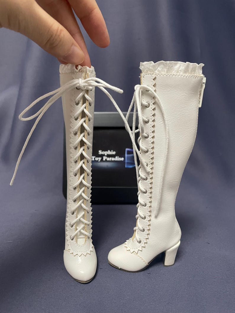 Pode incluir: Um par de botas brancas para bonecas com detalhes de atacadores e saltos pequenos. As botas t&ecirc;m uma borda ondulada na parte superior e s&atilde;o amarradas com atacadores brancos. As botas s&atilde;o projetadas para uma boneca.
