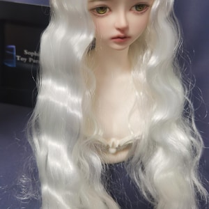 Könnte beinhalten: Ein weißer Puppenkopf mit langen, welligen, weißen Haaren. Die Puppe hat grüne Augen und einen neutralen Ausdruck.