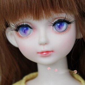 K&ouml;nnte beinhalten: Nahaufnahme eines Puppenkopfes mit langen braunen Haaren und blauen und violetten Iris. Die Puppe hat einen leichten rosa Rouge auf den Wangen und einen hellrosa Lippenstift.