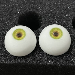 May include: Two white doll eyes with green irises and brown pupils. The eyes are on a black background.