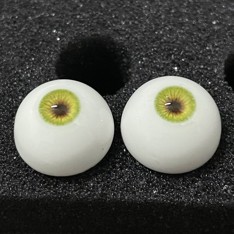 May include: Two white doll eyes with green irises and brown pupils. The eyes are on a black background.