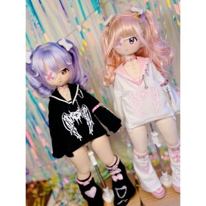 K&ouml;nnte beinhalten: Zwei Puppen mit rosa und lila Haaren, die schwarze und wei&szlig;e Outfits mit Engelsfl&uuml;geln tragen. Die Puppen tragen Augenklappen mit rosa Kreuzen. Die Puppen stehen auf St&auml;ndern.