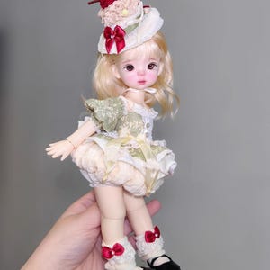 May include: A doll with blonde hair, wearing a green and cream-colored dress, cream-colored bloomers, and black shoes. The doll has a decorative hat with a red bow and a cherry on top. The doll is wearing white lace socks with red bows.