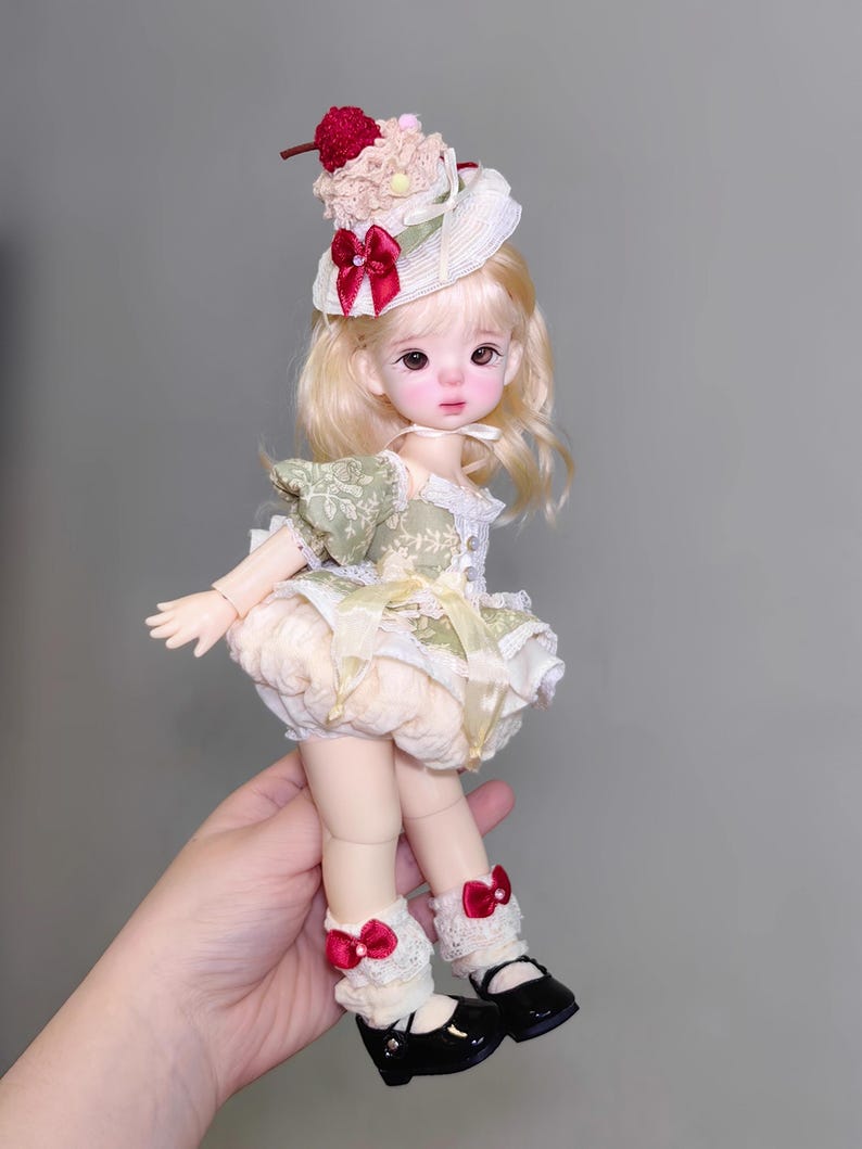 May include: A doll with blonde hair, wearing a green and cream-colored dress, cream-colored bloomers, and black shoes. The doll has a decorative hat with a red bow and a cherry on top. The doll is wearing white lace socks with red bows.