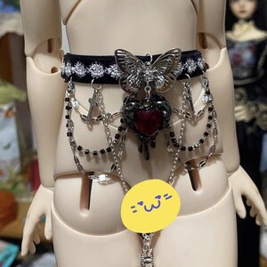 K&ouml;nnte beinhalten: Ein schwarzer Samt-Choker mit silbernen Ketten und einem baumelnden Herz-Anh&auml;nger. Das Herz tropft mit einer roten Fl&uuml;ssigkeit und hat einen silbernen Schmetterlings-Anh&auml;nger daran.