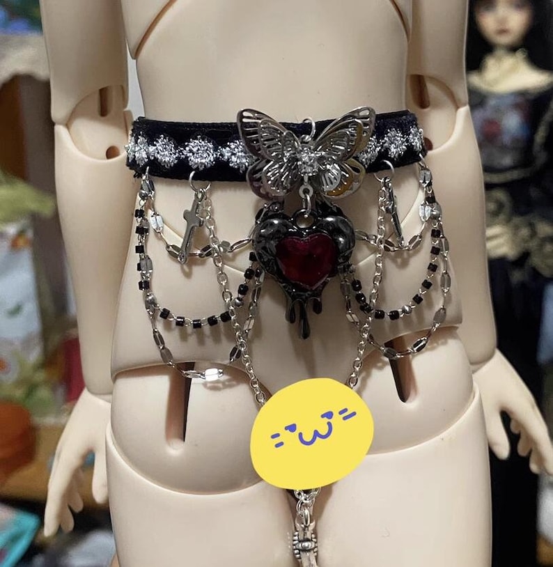 K&ouml;nnte beinhalten: Ein schwarzer Samt-Choker mit silbernen Ketten und einem baumelnden Herz-Anh&auml;nger. Das Herz tropft mit einer roten Fl&uuml;ssigkeit und hat einen silbernen Schmetterlings-Anh&auml;nger daran.