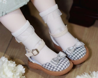 Può includere: Scarpe da bambola con motivo a quadretti e fiocco. Le scarpe hanno un cinturino beige con fibbia e sono abbinate a calzini bianchi con bordo in pizzo. La parola "Black" si trova nell'angolo in alto a destra.