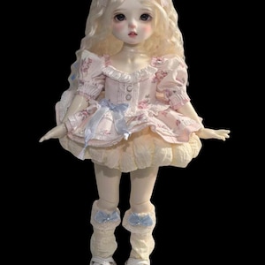 May include: A porcelain doll with long, wavy blonde hair and a pale complexion. The doll wears a pink floral dress with a ruffled skirt, blue bows, and white Mary Jane shoes. The doll's eyes are large and dark.
