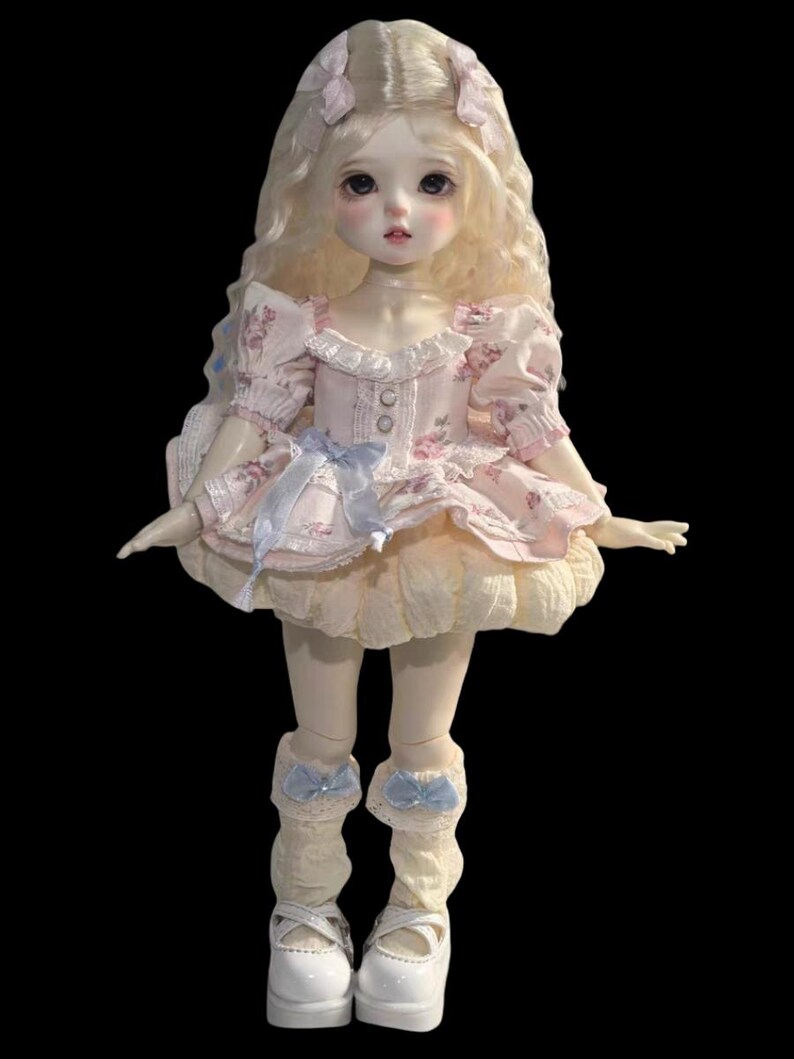May include: A porcelain doll with long, wavy blonde hair and a pale complexion. The doll wears a pink floral dress with a ruffled skirt, blue bows, and white Mary Jane shoes. The doll's eyes are large and dark.