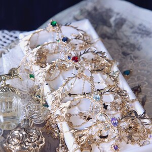 May include: A collection of gold-toned metal doll crowns featuring intricate branch-like designs, delicate chains, and various colorful faceted gemstones including red, blue, green, and purple. The text Only Doll Crown is visible at the top left.