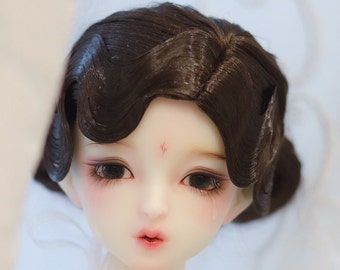 May include: A doll's face with dark brown, styled hair. The doll has large, dark eyes with long eyelashes, and a small red mark on its forehead. The doll's lips are a soft pink, and its skin is pale.