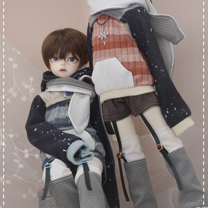May include: Two dolls wearing black and white patterned hooded sweaters with white accents. The dolls have brown hair and blue eyes. One doll is wearing a plaid shirt underneath the sweater. The dolls are standing on a light pink background.