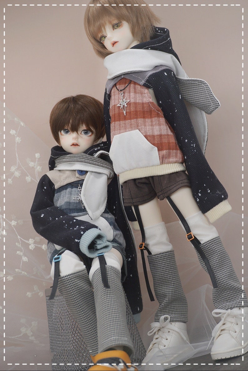 May include: Two dolls wearing black and white patterned hooded sweaters with white accents. The dolls have brown hair and blue eyes. One doll is wearing a plaid shirt underneath the sweater. The dolls are standing on a light pink background.