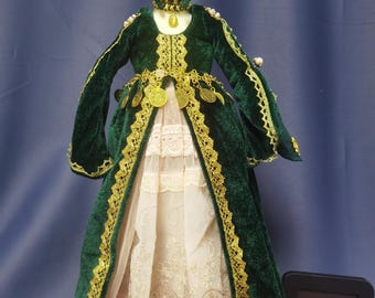 May include: A green velvet doll dress with gold trim and a white lace underskirt. The doll is wearing a gold crown with a star on top. The doll is standing on a wooden base. The image also includes a small black sign with the text "Sophie Toy Paradise".