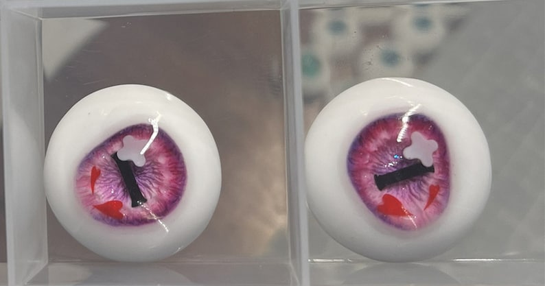 May include: Two white doll eyes with a purple and pink iris, a black pupil, and white and red accents.