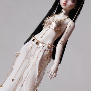 May include: A porcelain doll with long black hair wearing a gold and white costume with a star-shaped headpiece. The doll is wearing a gold chain belt with dangling coins and a gold chain bra with a mesh design.