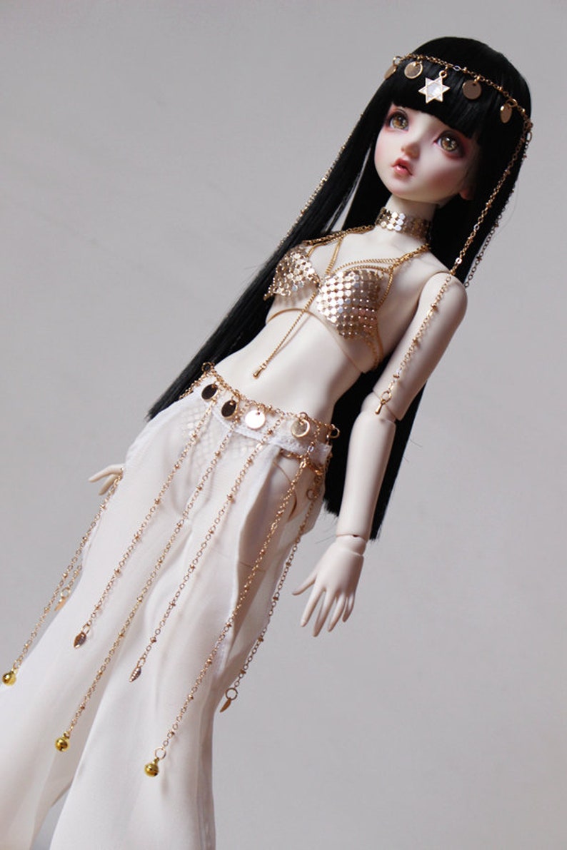 May include: A porcelain doll with long black hair wearing a gold and white costume with a star-shaped headpiece. The doll is wearing a gold chain belt with dangling coins and a gold chain bra with a mesh design.