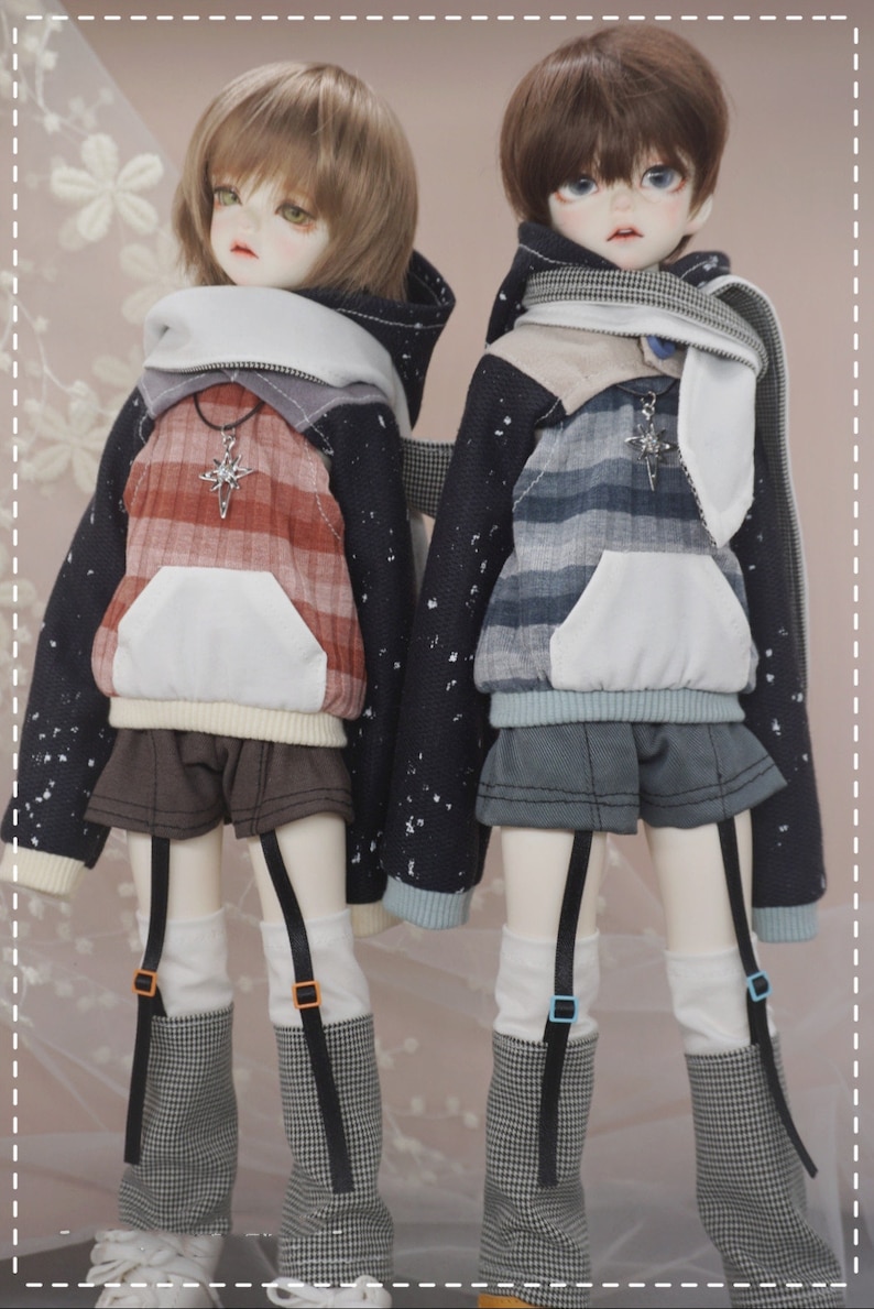 May include: Two dolls wearing black and white patterned hoodies with white collars and matching black and white checkered leggings. The doll on the left is wearing a red and white plaid shirt underneath the hoodie and the doll on the right is wearing a blue and white plaid shirt underneath the hoodie.