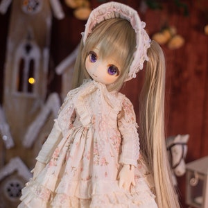 May include: A doll with long blonde hair wearing a white dress with pink floral print and a white bonnet. The doll has purple eyes and is standing in front of a wooden background.