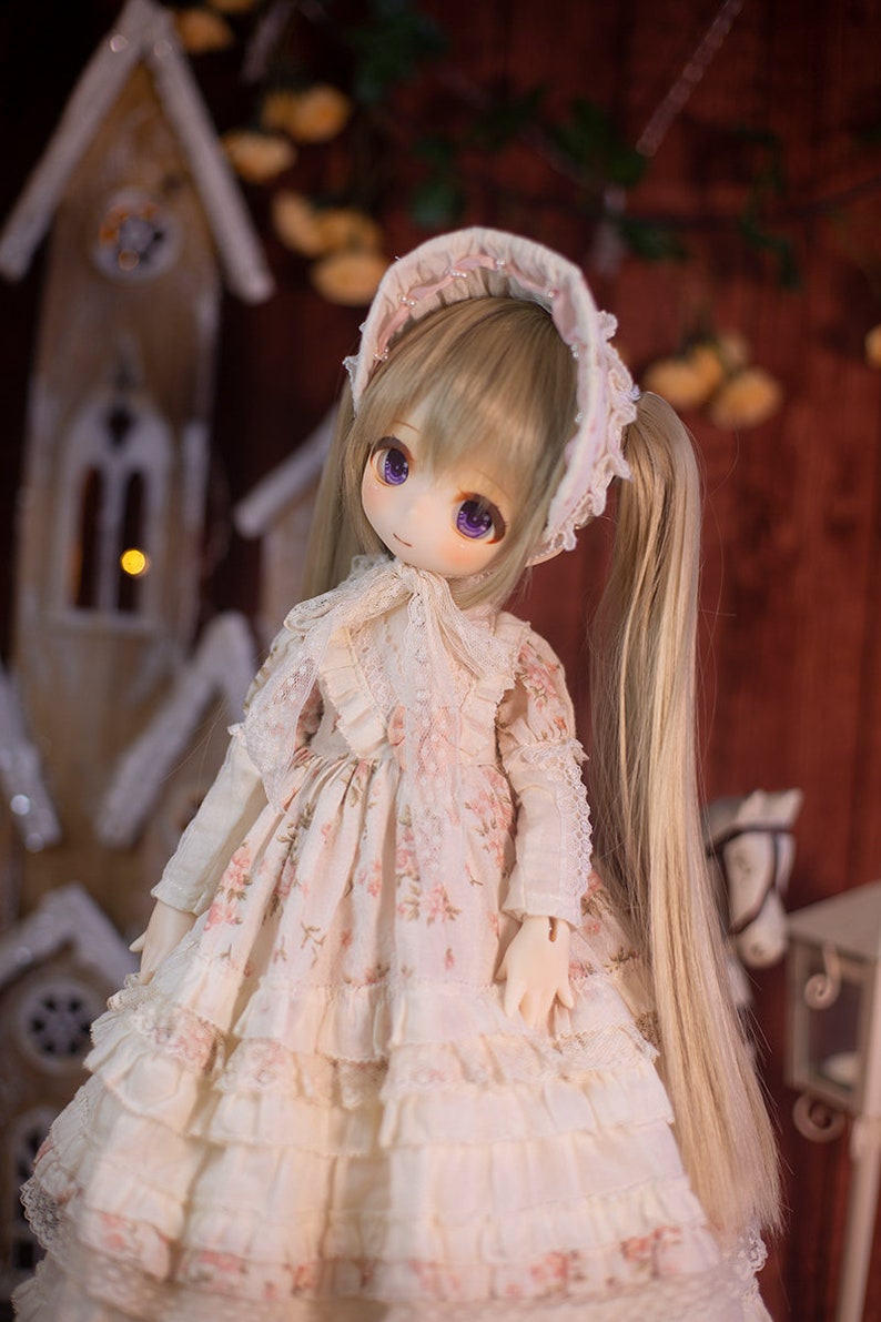 May include: A doll with long blonde hair wearing a white dress with pink floral print and a white bonnet. The doll has purple eyes and is standing in front of a wooden background.