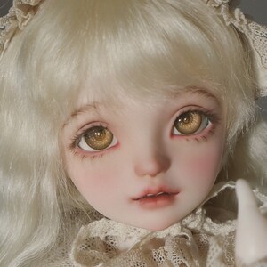 Puede incluir: Una mu&ntilde;eca de porcelana con cabello rubio y ojos marrones que lleva un vestido de encaje blanco.