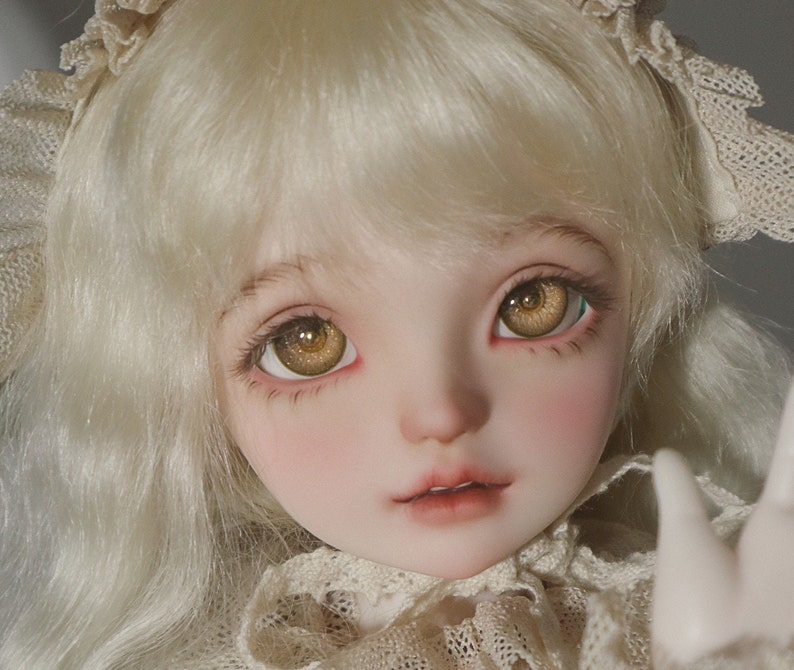 Puede incluir: Una mu&ntilde;eca de porcelana con cabello rubio y ojos marrones que lleva un vestido de encaje blanco.
