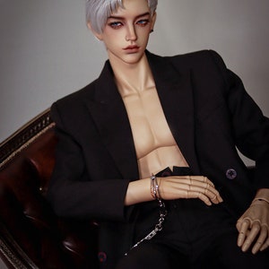 May include: A doll wearing a black suit jacket and black pants. The doll has white hair and blue eyes. The doll is wearing silver handcuffs on one wrist.