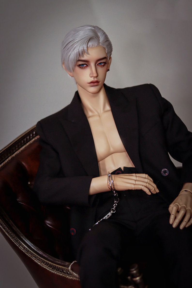 May include: A doll wearing a black suit jacket and black pants. The doll has white hair and blue eyes. The doll is wearing silver handcuffs on one wrist.
