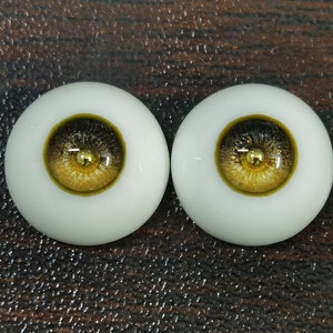 Puede incluir: Dos ojos de mu&ntilde;eca realistas con escler&oacute;tica blanca, iris negro y pupila dorada y marr&oacute;n. Los ojos est&aacute;n colocados sobre un fondo marr&oacute;n.